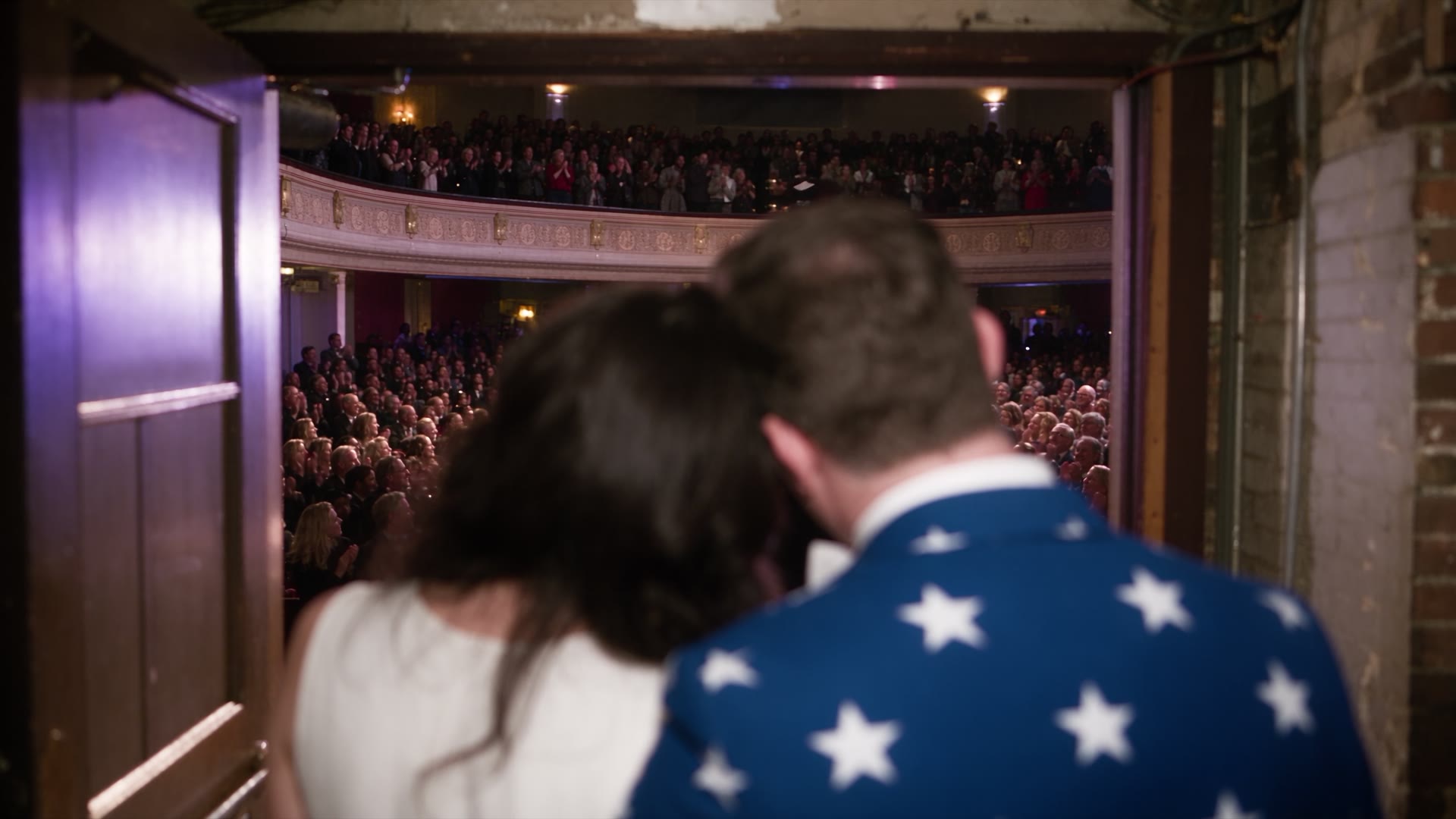 Seen from the wings, a couple leans together as a packed theater rises in applause. He wears a navy jacket covered in white stars.